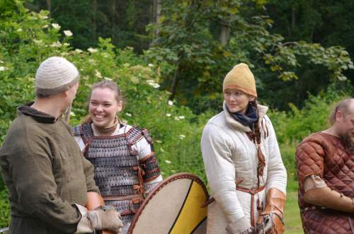 Photo: Die Äußerungen eines Wikingers mit nadelgebundener Mütze führen in dieser Runde zur Erheiterung der Kämpferinnen. Im Hintergrund blühen die Sträucher und  Bäume stehen in sattem Grün.